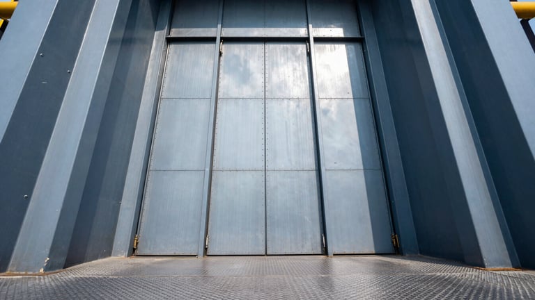 Low-angle architectural shot of a massive industrial freight elevator with open heavy steel doors, rugged textured steel floors, cool high-contrast daylight.