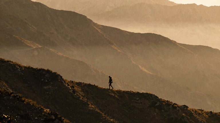 Wide environmental landscape of a solitary figure walking along a misty mountain ridge at golden hour, deep charcoal shadows and warm amber light, 35mm grain.