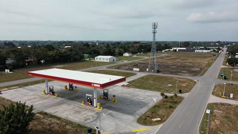 Wide drone overhead shot, overcast daylight, a completed fuel station canopy and pump islands at left, a three-legged cell tower visible in the mid-ground right, open lot and access road showing full site footprint and landscape relationship