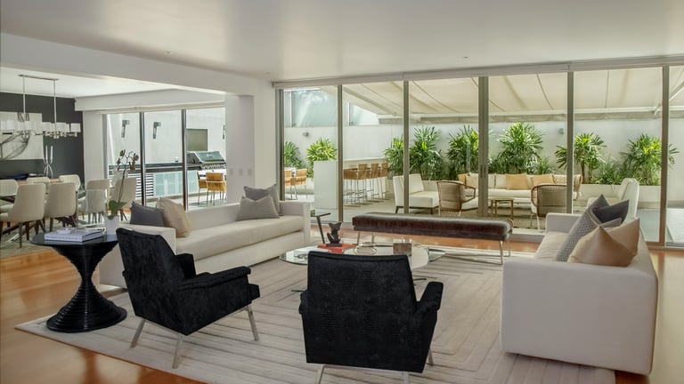 Modern luxury living room with white sofas, black velvet chairs, and sliding glass doors leading to a patio.