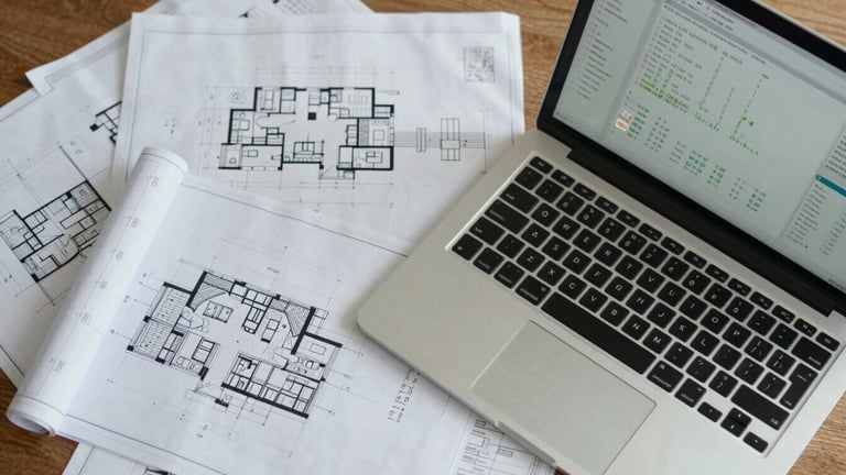 Top-down structured desk layout showing architectural blueprints next to a modern laptop screen displaying data charts, crisp daylight.