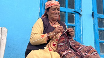 Woman in traditional dress sitting outside blue home and knitting a blanket.