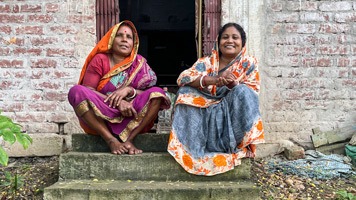 2 women in sarees sitting on step In front of building..