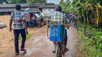 2 men  one walking and one cycling, on narrow gravel road approaching school.