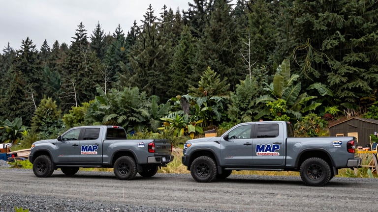 Wide shot of two branded MAP Engineering Inc. pickup trucks parked on a gravel road at a project site, lush Oregon forest in background, overcast natural daylight, cool slate and forest green tones, sharp focus, 35mm, documentary engineering photography.