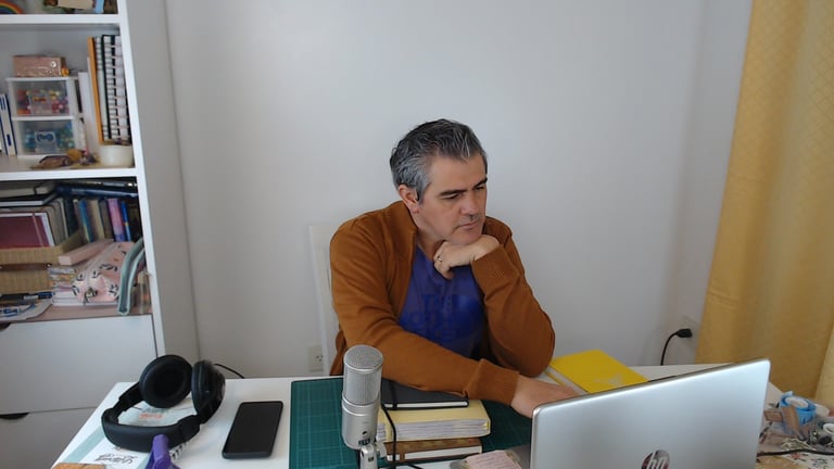 A man in a brown cardigan working on his HP laptop at a white desk with a microphone and headphones.