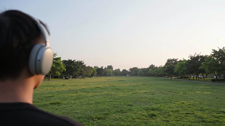 Wide panoramic view of a serene park at sunrise, a person in the foreground enjoying the view with the open-ear headset on, expansive sky, low horizon, soft morning light.