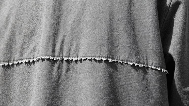 Extreme macro crop of raw-edged hem stitchwork on a heavy-gauge canvas garment, cold overcast daylight, sharp focus, brutalist aesthetic.