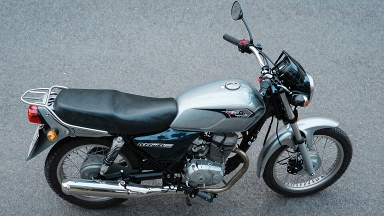 Overhead studio shot of a 150cc commuter motorcycle laid flat on a cool grey surface, instrument console and fuel tank badge in sharp focus, even neutral daylight from above revealing seat stitching and panel geometry