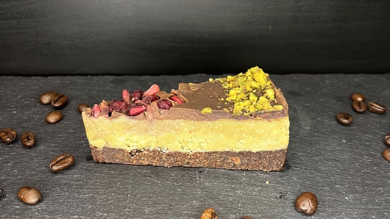 A slice of vegan raw cake with pistachio and dried berries on a slate board with roasted coffee beans.