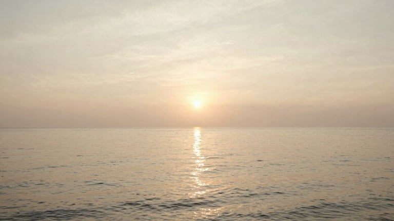 Wide panoramic golden hour shot over Southeast Asian coastline, soft warm sunlight reflecting on calm water, low horizon, serene atmosphere, 35mm film style.
