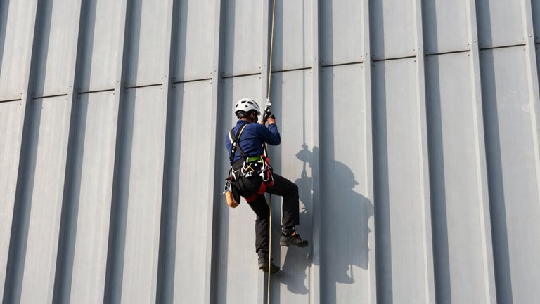 Rope-access technician in full harness and helmet working on a vertical steel industrial facade, rigging lines taut, hands gripping a tool at a structural connection point, hard daylight shadows on the structure surface, wide framing showing height and scale