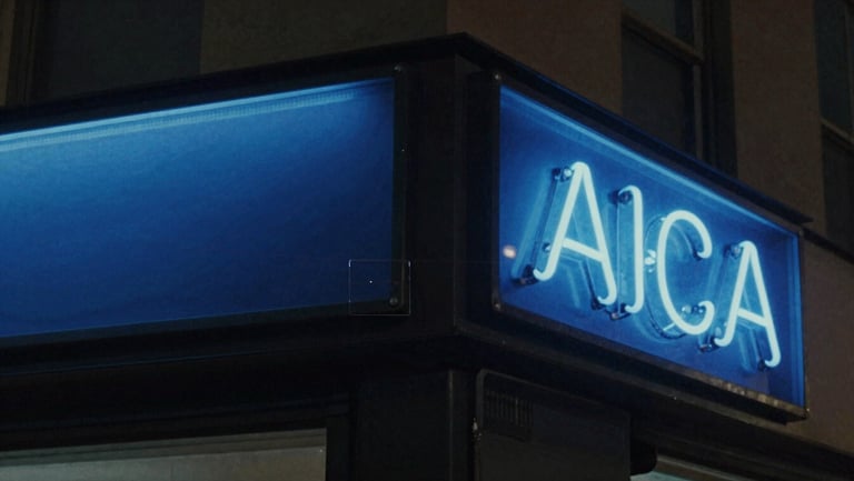 Analog video still with visible scanlines of a dark street corner at night, glowing blue neon sign, grainy texture.