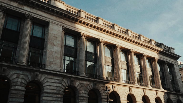 Low-key architectural shot of a historic London neoclassical facade blended with modern glass, dramatic chiaroscuro lighting, deep obsidian and gold tones, 35mm film grain.
