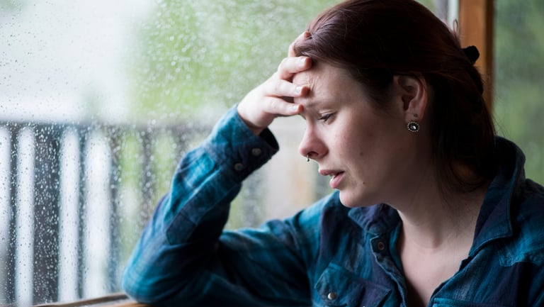A stressed woman rests her head in her hand by a rainy window, depicting anxiety and mental health struggles.