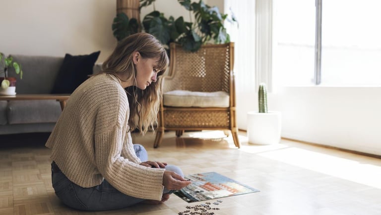 A woman sits on a wooden floor in a sunny living room assembling a jigsaw puzzle.