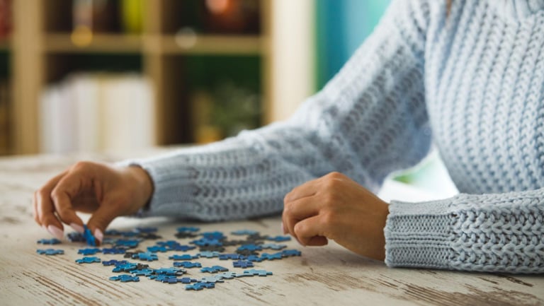 A woman in a blue sweater assembles a blue jigsaw puzzle on a rustic wooden table.