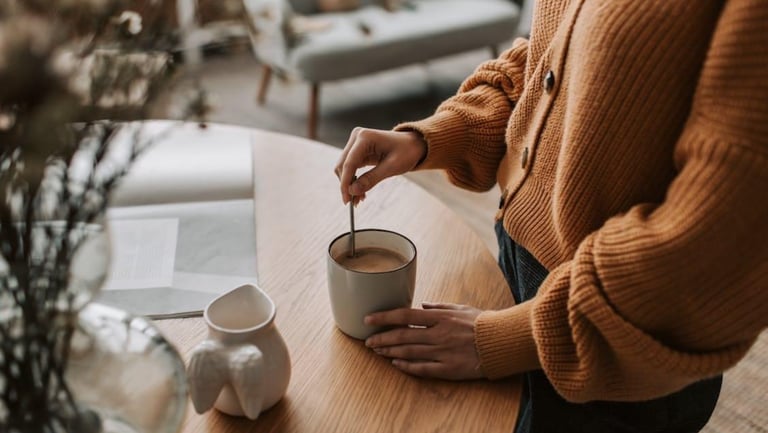 A person in a brown knit sweater stirs a cup of coffee on a wooden table.