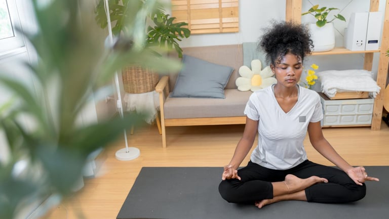 A young woman practicing yoga meditation in a lotus pose on a mat in her bright living room.