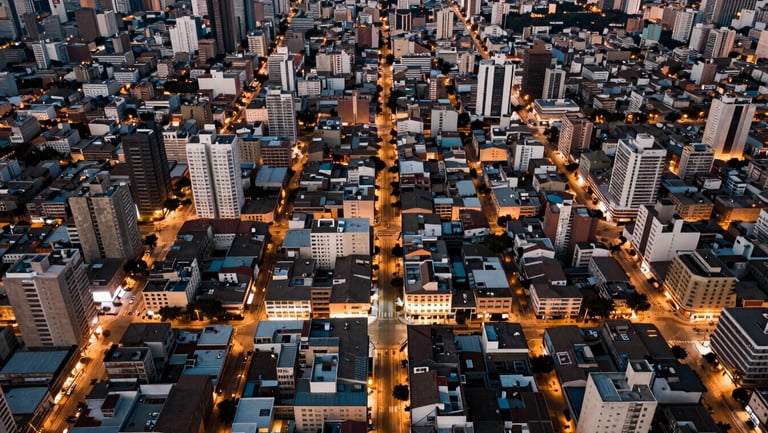 Bird's-eye elevated perspective of a dense Lima urban neighborhood at dusk, city grid stretching to the horizon, warm amber streetlights beginning to illuminate, no people, architectural scale and urban density as the subject