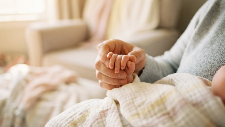 A close-up of a parent gently holding a baby's tiny hand, soft warm morning window light, pastel blankets, shallow depth of field, 35mm film.