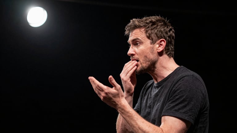 Close environmental frame, actor mid-scene under a single hard spotlight in an otherwise black theatre, face three-quarter turned, tension in jaw and hands, documentary capture, high contrast