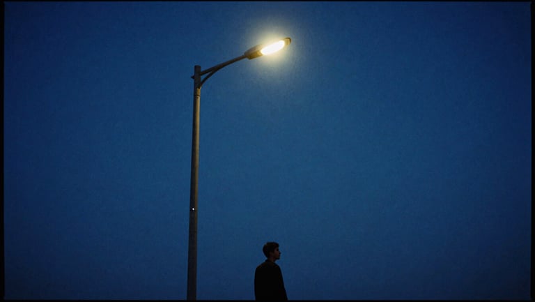 Anamorphic film still of a solitary figure standing under a single streetlamp at twilight, cool blue moonlight, high contrast chiaroscuro