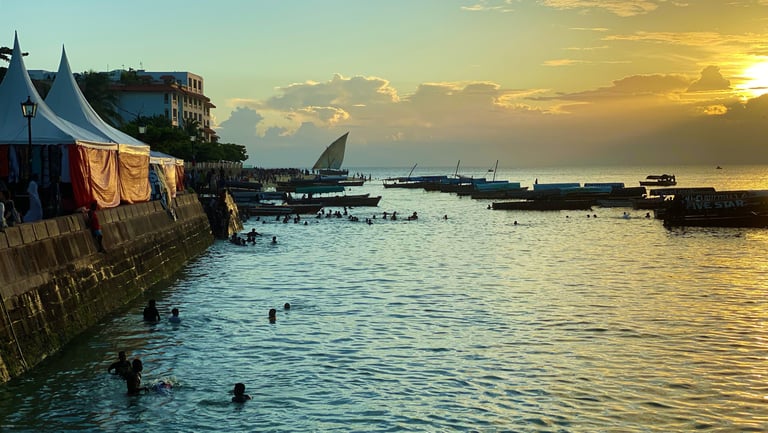 Sunset over a stone seawall with people swimming and a traditional dhow on the water.