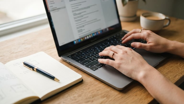 Close-up of hands typing on a laptop on a clean wooden desk under soft natural window light, an open notebook with gold pen nearby, 35mm editorial photography
