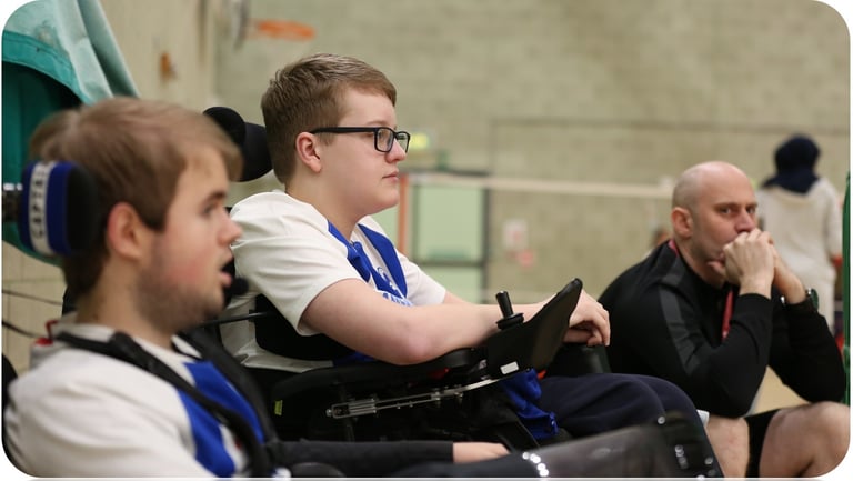 Indoor sports setting with young powerchair football players sitting side-by-side and watching