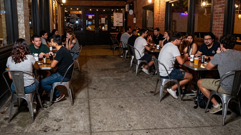 A wide-angle direct flash photograph of the concrete sidewalk outside the taproom at night, simple metal tables crowded with people, cozy chaotic arts district energy