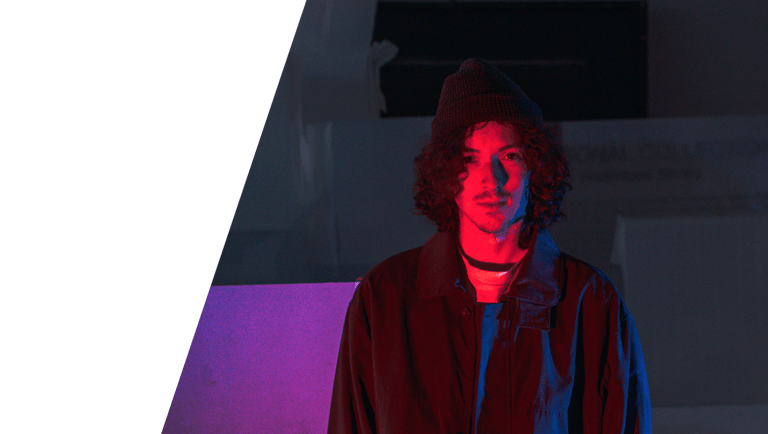 A dark portrait photographic image of In Videcolour lead-songwriter Sam Auckland. Sam is standing, red and blue light on him.