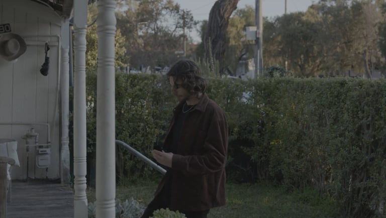 A man with long hair wearing a brown jacket stands on a porch next to a white pillar and green hedge