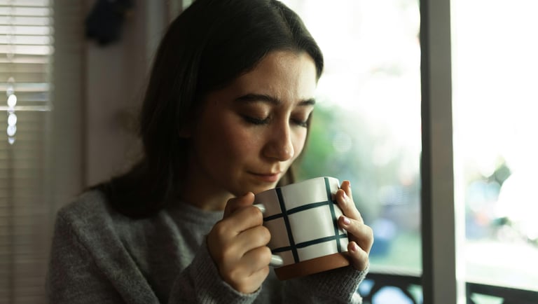 A young woman in a grey sweater holds a grid pattern coffee mug while enjoying a morning drink.