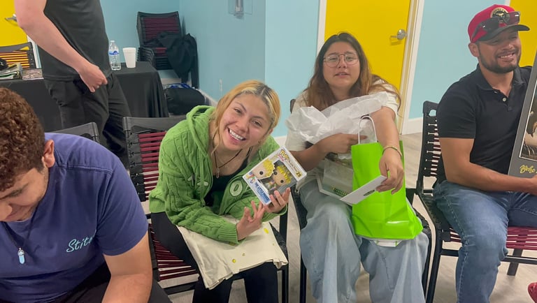 Two smiling women sitting indoors holding a Funko Pop The Little Prince collectible and a green gift bag.