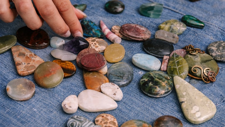 Mano seleccionando piedras naturales pulidas y minerales semipreciosos sobre tela de mezclilla azul para joyería artesanal.