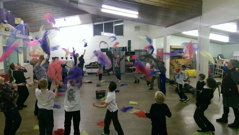 Children and adults throwing colourful juggling scarves in a vibrant indoor sensory workshop.
