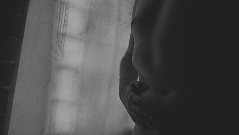 Fotografía desnudo artístico hombre con mano sobre el abdomen frente a cortina floral.