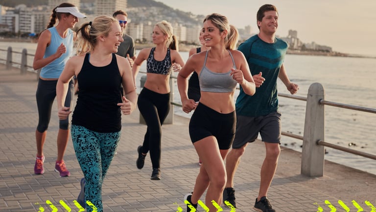 A diverse group of friends in athletic wear jogging together on a sunny coastal promenade at sunset.