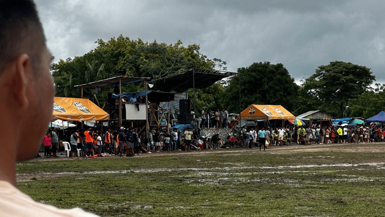 Crowd gathered at a muddy outdoor festival with tents and a stage under a cloudy sky.