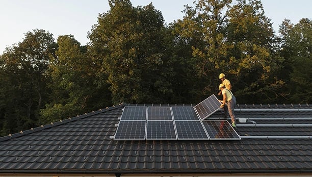 Professional technicians installing residential solar panels on a dark tiled roof near a forest.