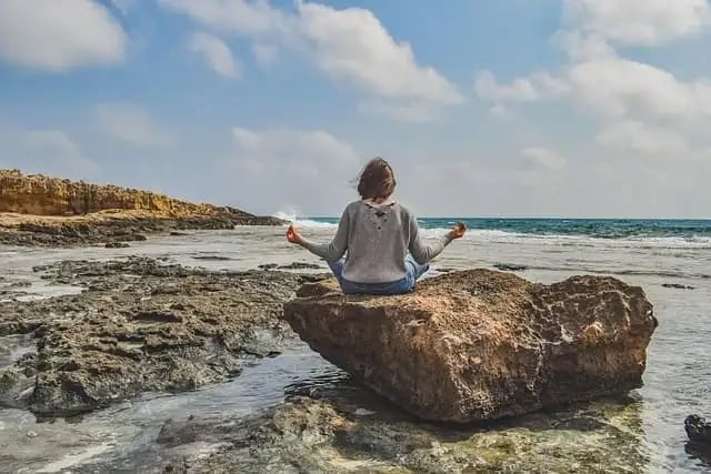 Femme assise sur un rocher face à la mer, mains écartées, doigts joints en posture de méditation