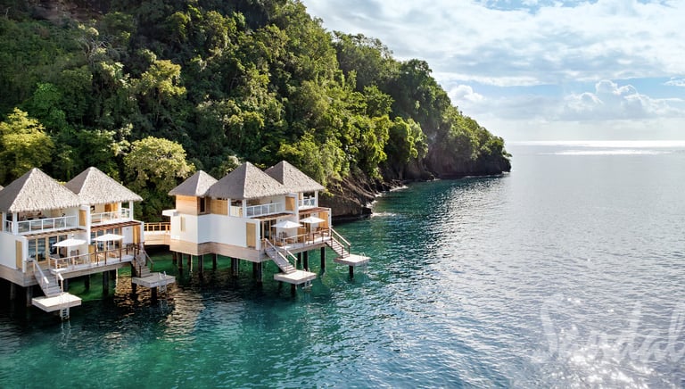 Luxury overwater bungalows at a tropical resort nestled against a lush green cliffside by the ocean.