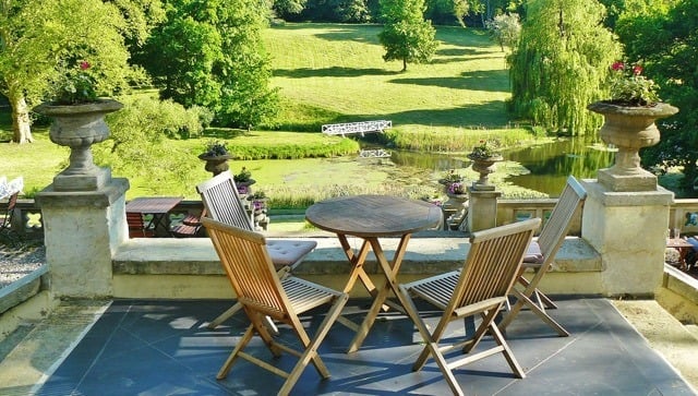 acid stained stamped concrete patio with wooden furniture overlooking green property with trees, a small bridge and a lake