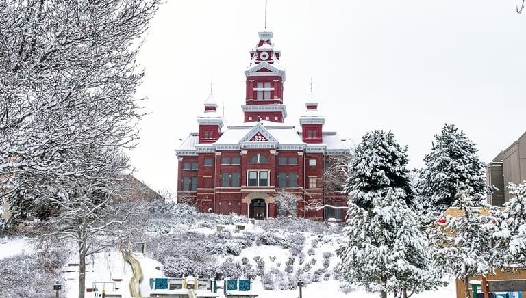 Historic red brick clock tower building standing behind a snow-covered winter park and playground.