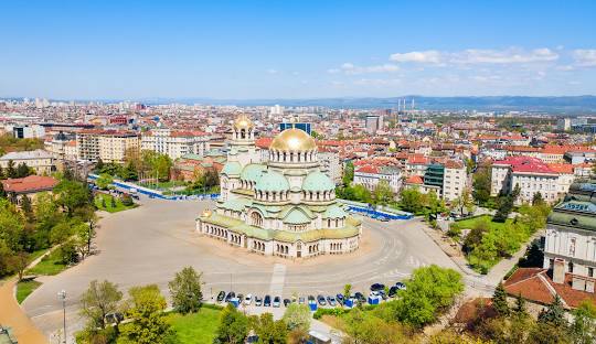 A sunny view of Vitosha Boulevard in Sofia, Bulgaria, featuring a tree-lined pedestrian street and mountain views.