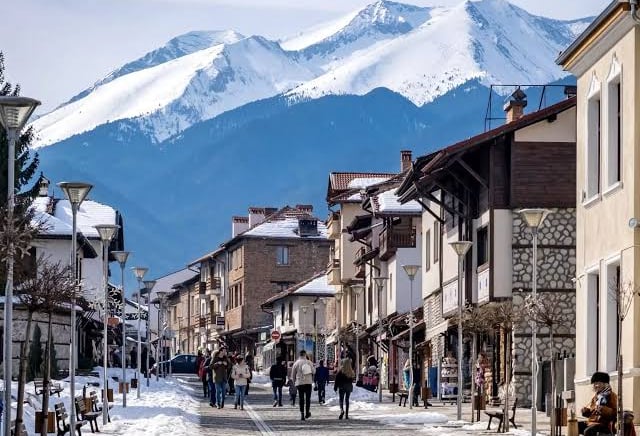 Tourists walking through the snowy streets of Bansko ski resort town with Pirin Mountains in the background.