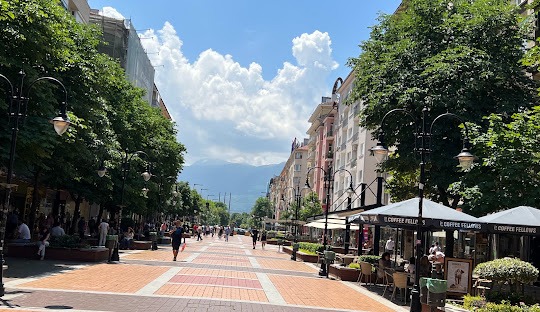 Sunny view of Vitosha Boulevard in Sofia, Bulgaria, featuring outdoor cafes, lush trees, and a mountain backdrop.