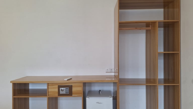Modern hotel room furniture featuring a wooden desk with a digital safe, mini fridge, and open wardrobe shelving.