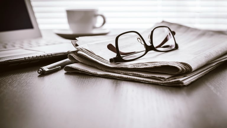 A desk with a laptop, a pen, a cup of tea, and a newspaper with a pair of reading glasses on it.